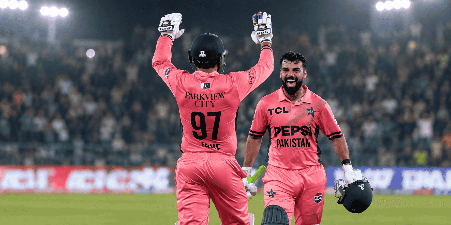 Two Pakistani cricket players in pink uniforms celebrate a wicket under stadium floodlights, with